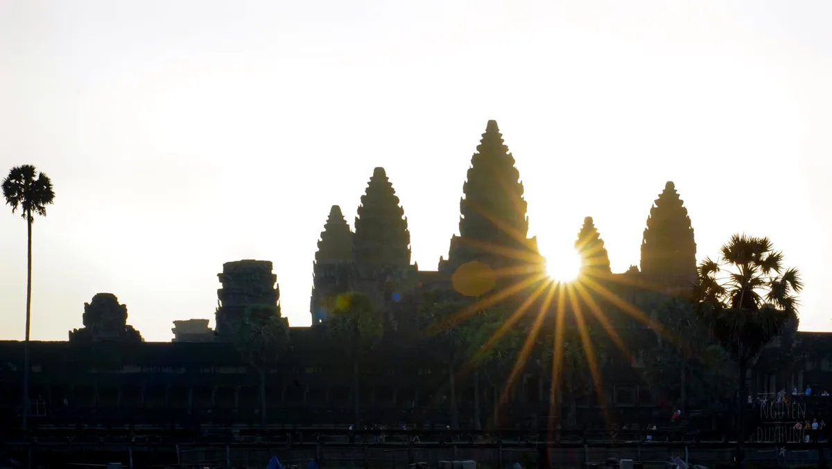 The sun rising directly behind the historic stone spires of Angkor Wat, casting perfect reflections across the lotus pond.